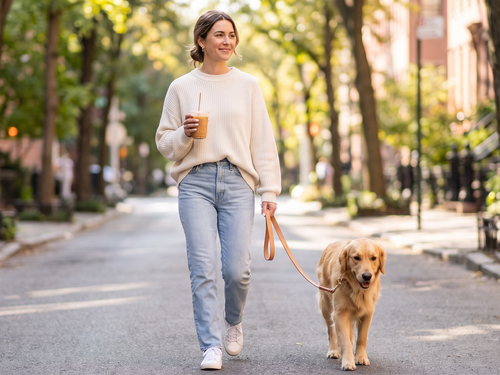 Woman walking a golden retriever on a sunny sidewalk, holding an iced coffee, wearing pink almond nails