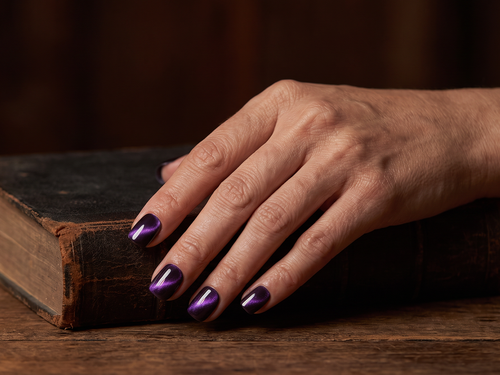 Hand wearing deep violet cat eye square press-on nails resting on an old leather book beside a lit candle