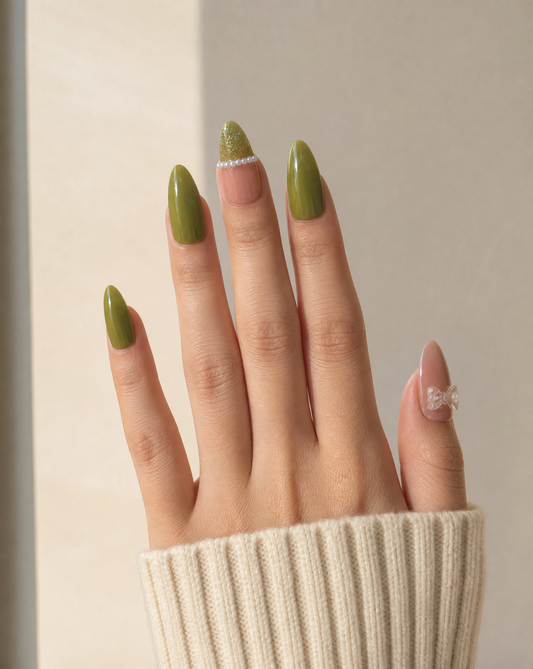 Hand in a cream knit sleeve wearing long olive green almond press-ons with a pearl line and a clear bow thumb