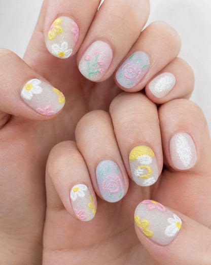Hands wearing milky pastel square press-ons with textured daisies, roses, and a yellow moon