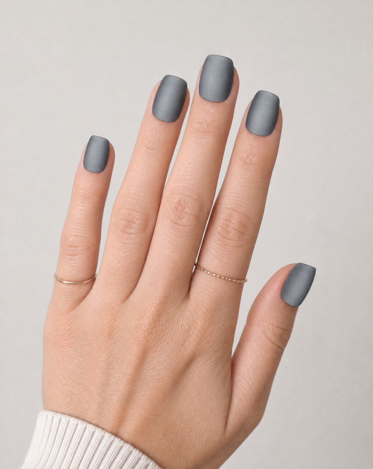 Hand with thin gold rings wearing matte charcoal gray square press-ons
