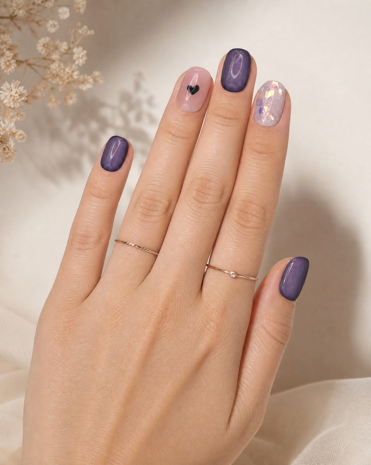 Hand with thin gold rings wearing plum square press-ons with a black heart and a glitter nail