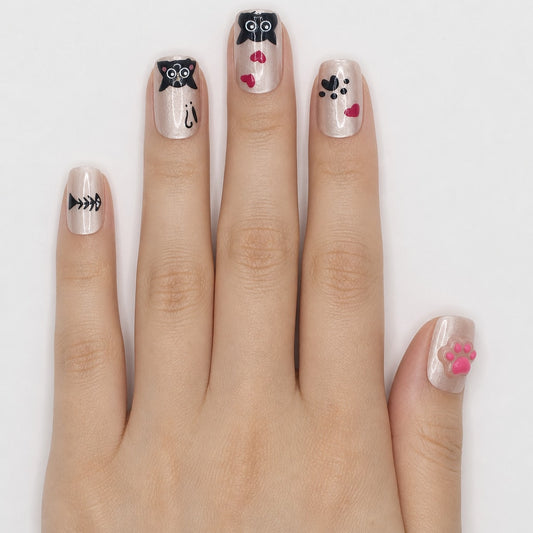 Hand wearing champagne square press-ons with black kawaii cats, a pink 3D paw, and fish bones