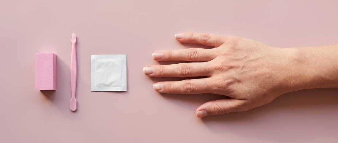 Relaxed bare hand with short natural nails beside a buffing block, cuticle pusher, and alcohol pad on a blush pink surface