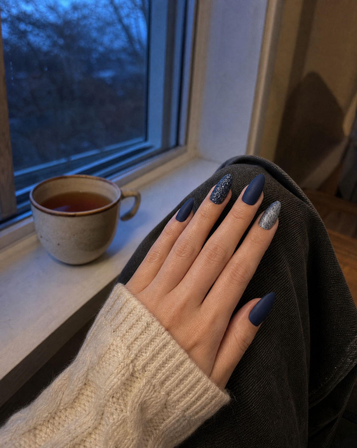 Hand with matte midnight navy almond press-on nails resting on a knee by a window at dusk, a cup of tea on the sill